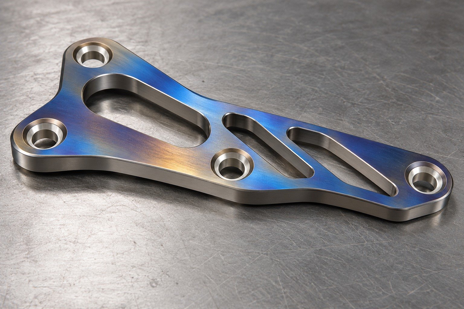Distinct titanium asymmetric slotted plate with muted blue-gold anodized appearance on broad face and bright masked mounting hole surfaces,illustrative appearance only. — illustrative generated component concept